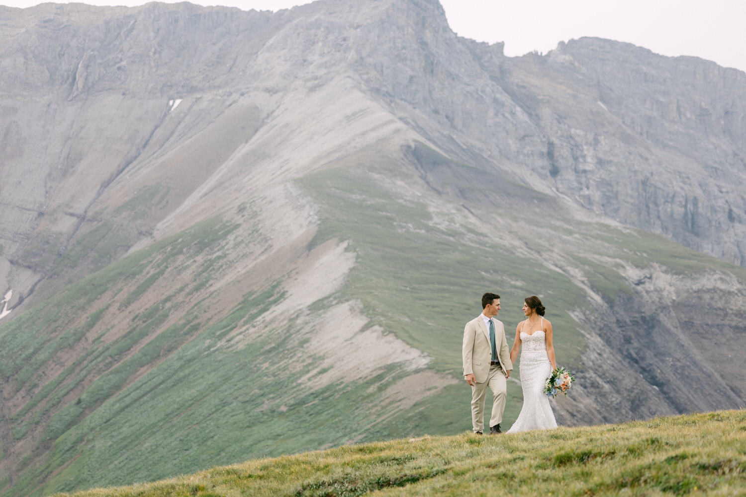 A couple strolls hand in hand on a grassy hilltop, surrounded by stunning mountain scenery, capturing a moment of joy and love on their special day.