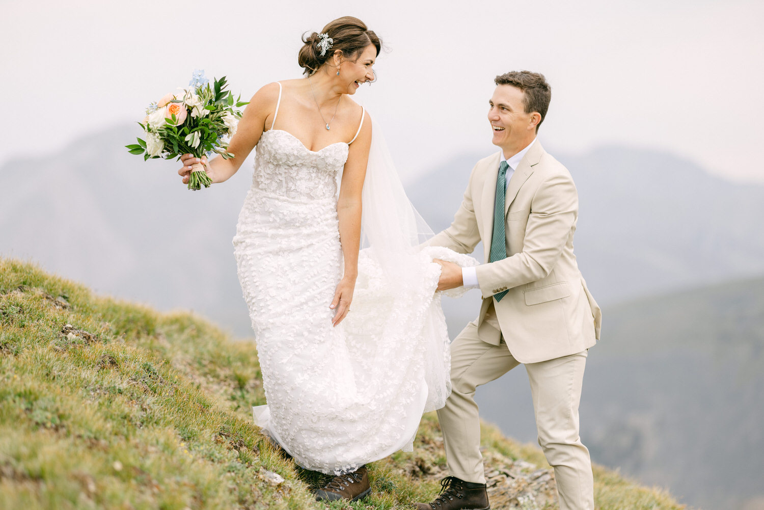 A bride and groom share a joyful moment on a hillside, with the bride holding a bouquet and the groom offering support while lifting her dress.