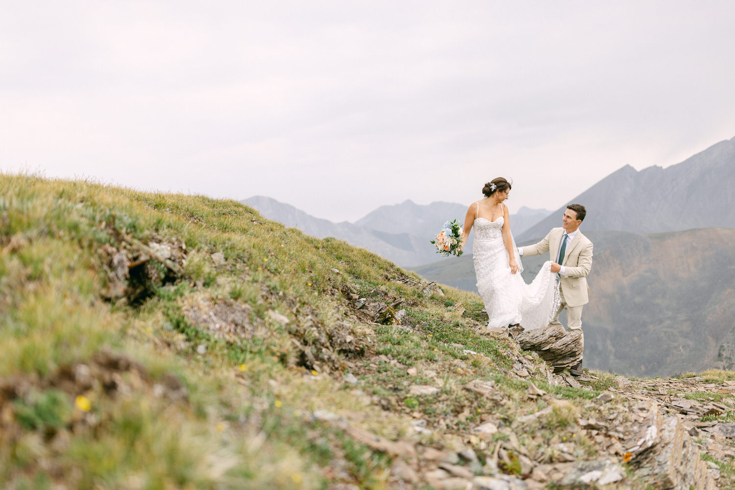 A bride in a white gown and groom in a beige suit share a romantic moment on a rocky hillside surrounded by mountains and greenery.