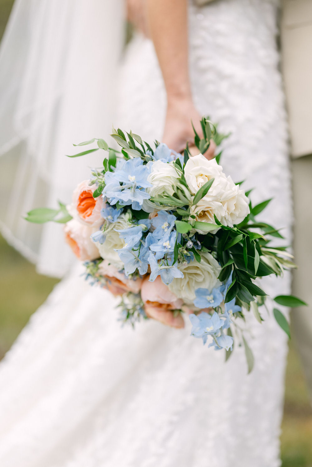 A close-up of a bride holding a lush bouquet filled with pastel flowers including blue delphiniums, peach roses, and greenery, set against her textured white wedding dress.