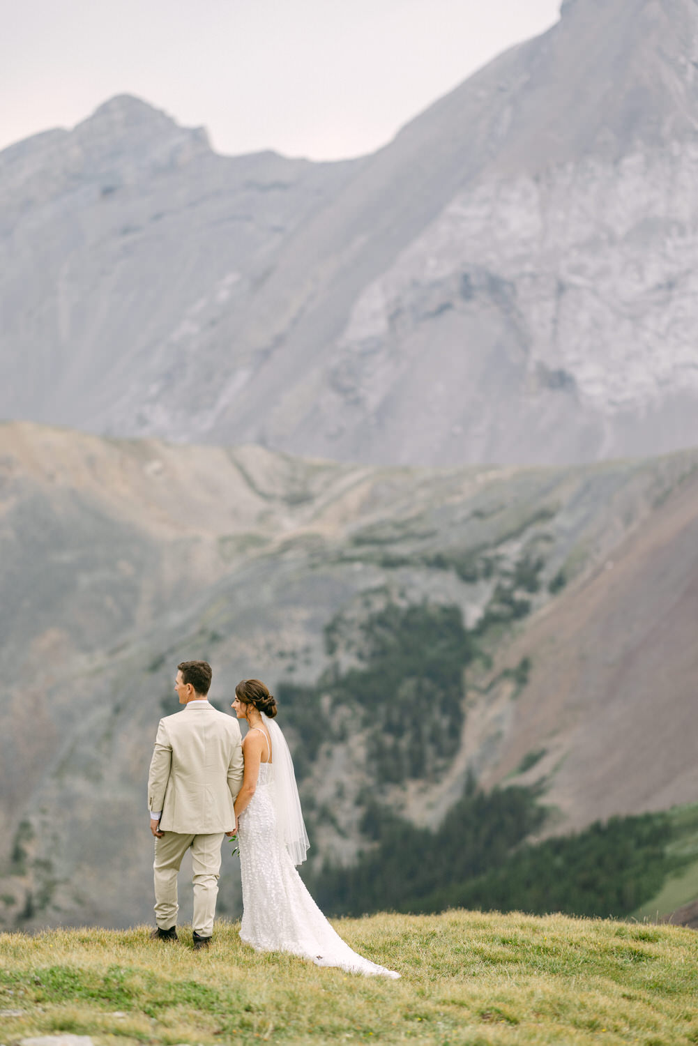 A couple holds hands in a stunning mountainous landscape, dressed in wedding attire, captured from behind.