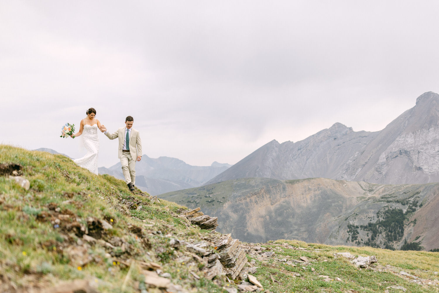 A couple in wedding attire walks hand-in-hand down a grassy slope, surrounded by mountains and cloudy skies.