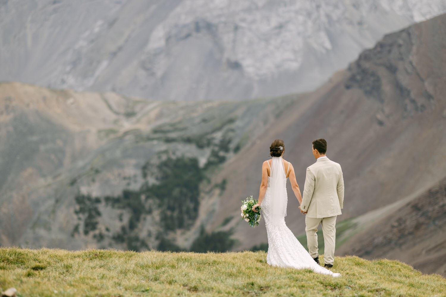 A bride and groom holding hands, standing on a grassy ledge with a stunning mountain backdrop, capturing a moment of tranquility and love.