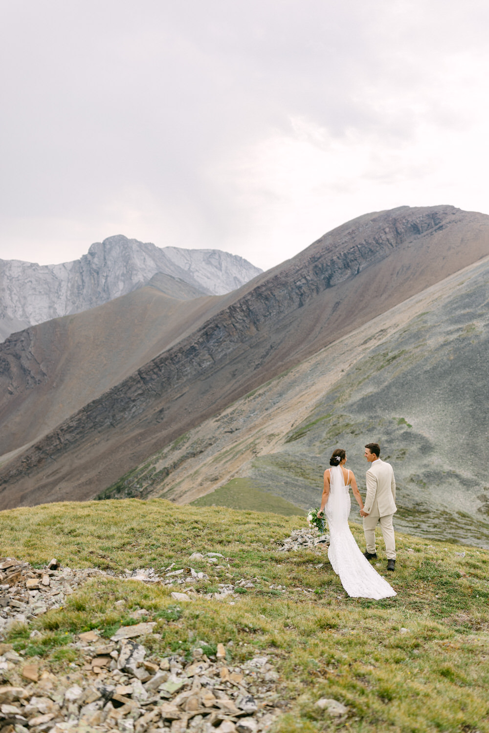 A couple in wedding attire stands hand-in-hand on a grassy hill, overlooking dramatic mountain scenery.