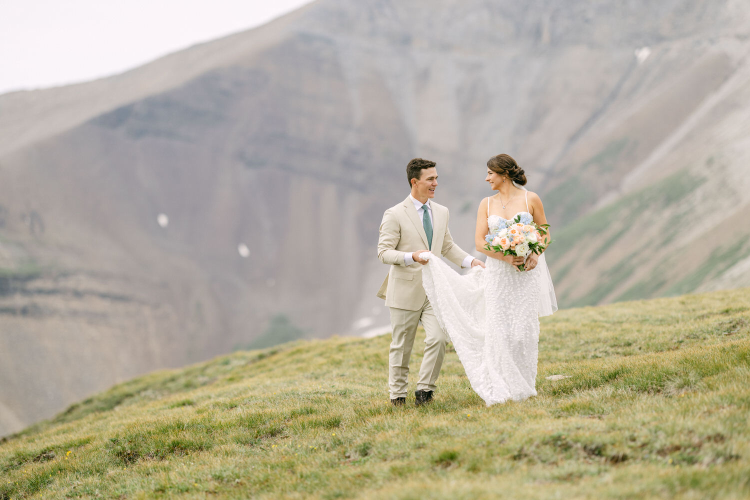 A couple walks hand-in-hand on a grassy hillside surrounded by mountains, with the bride holding a bouquet and wearing a flowing white dress, and the groom in a light suit.