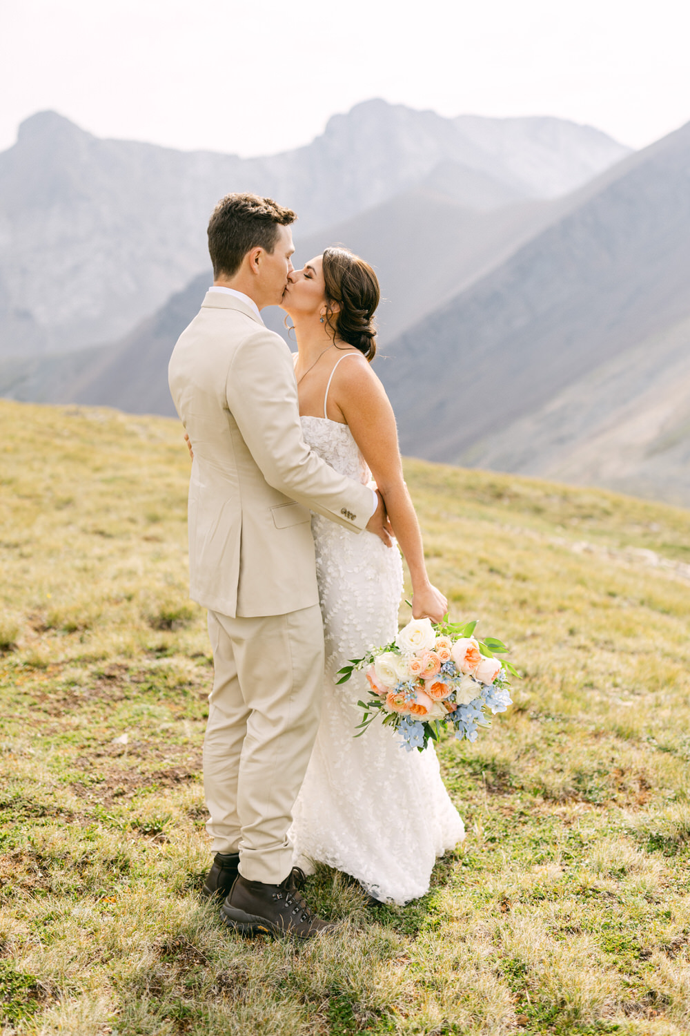 A bride and groom share a kiss amidst a picturesque mountain backdrop, with the bride holding a vibrant bouquet of flowers.