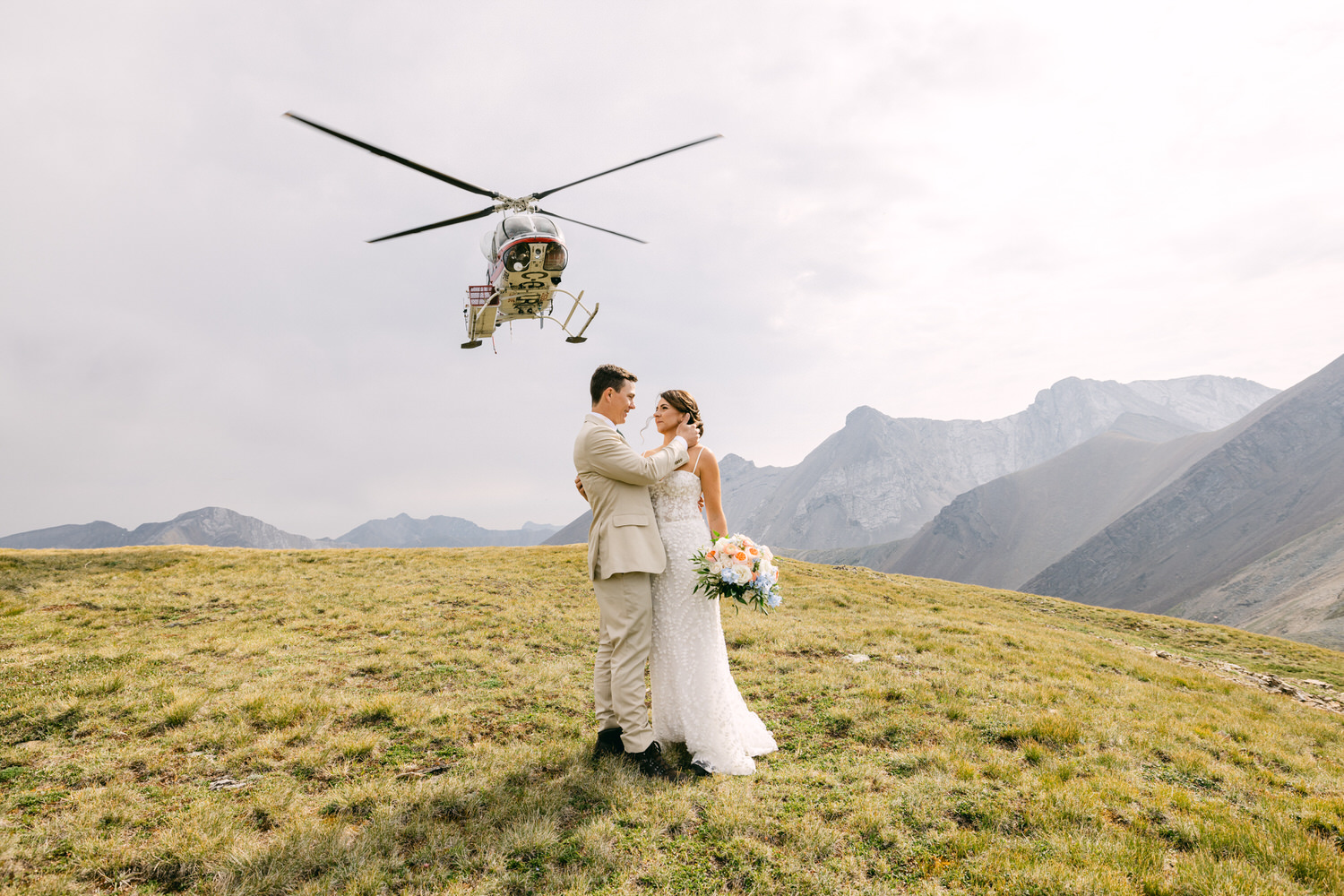 A bride and groom embrace on a grassy mountain ridge with a helicopter flying overhead, set against a backdrop of dramatic peaks and cloudy skies.