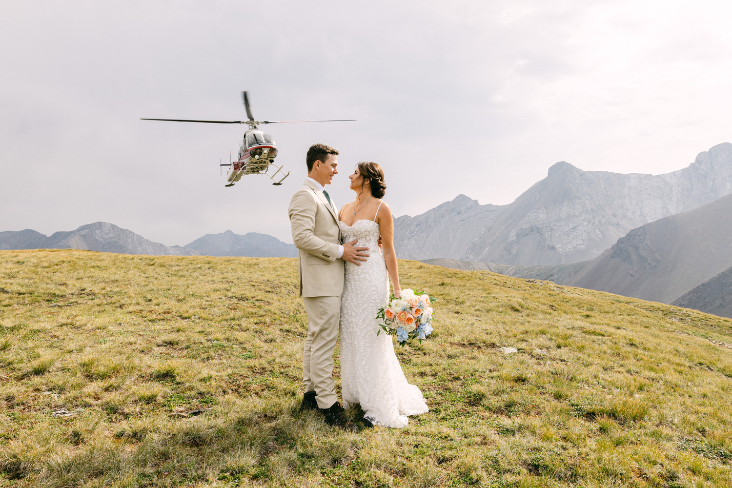 A couple embraces on a grassy hilltop, with a helicopter flying above and majestic mountains in the background.