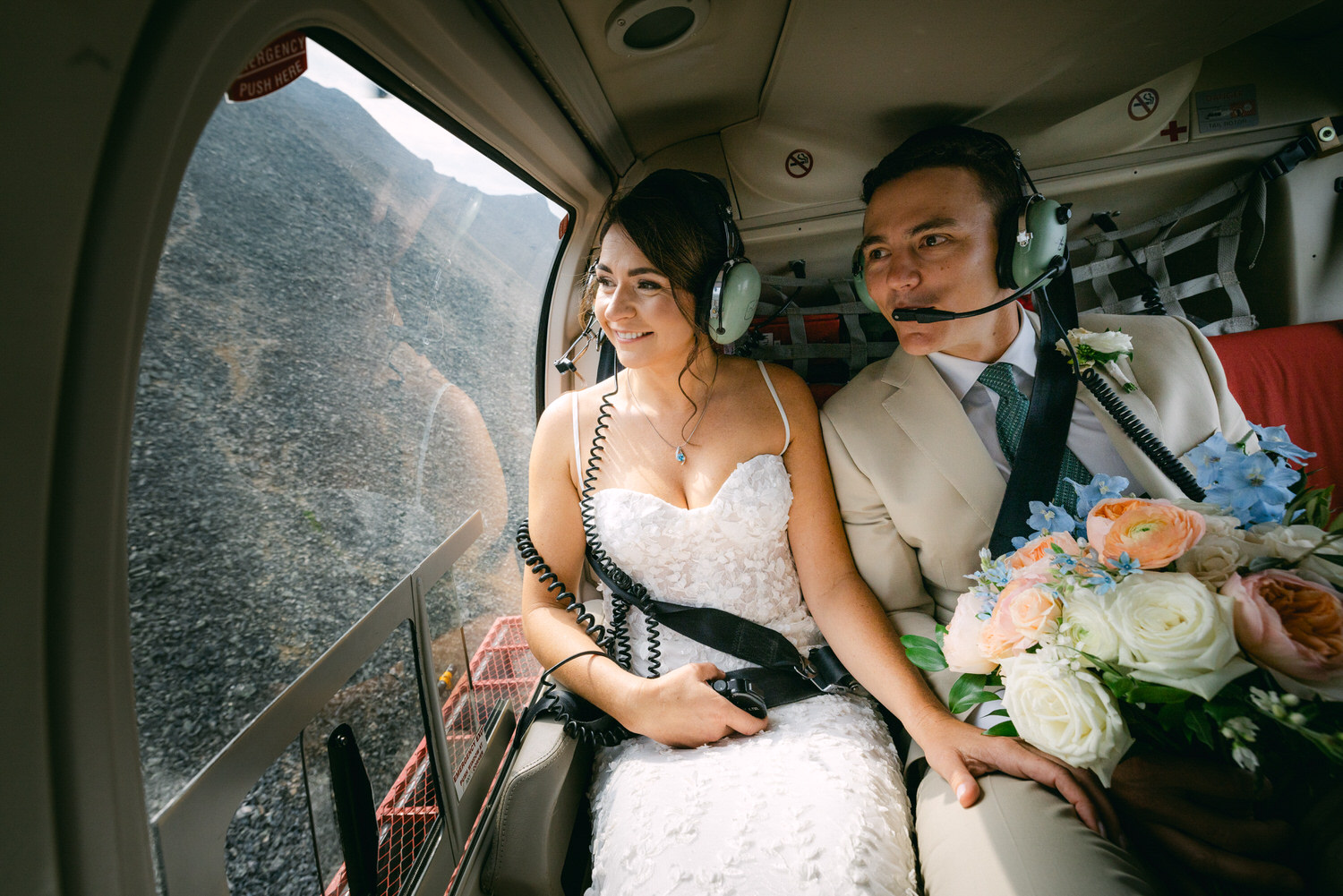 A bride and groom wearing headsets smile together while seated in a helicopter, admiring the rocky landscape outside.