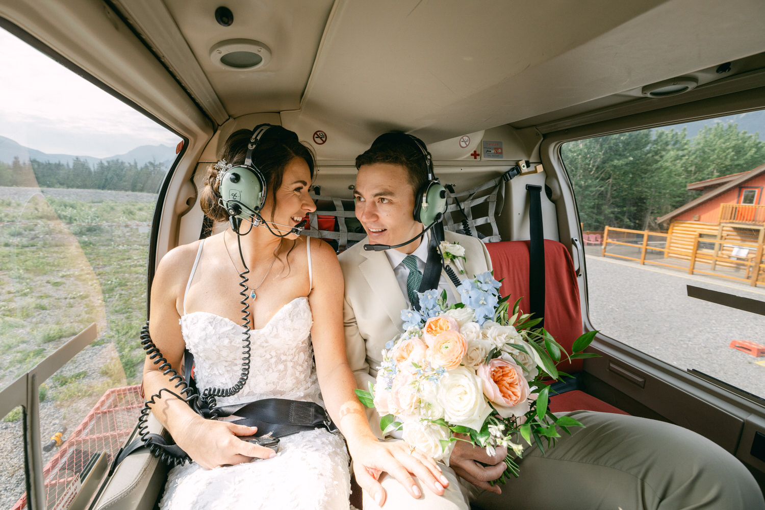 A bride and groom in a helicopter, wearing headsets, sharing a joyful moment as they prepare for their wedding flight, with a beautiful bouquet in the bride's hands.