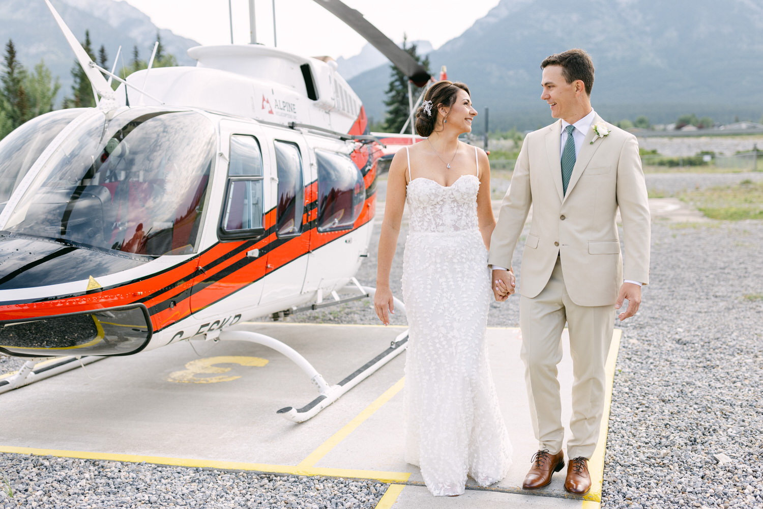 A joyful couple walks hand in hand near a helicopter, dressed in wedding attire against a stunning mountain backdrop.