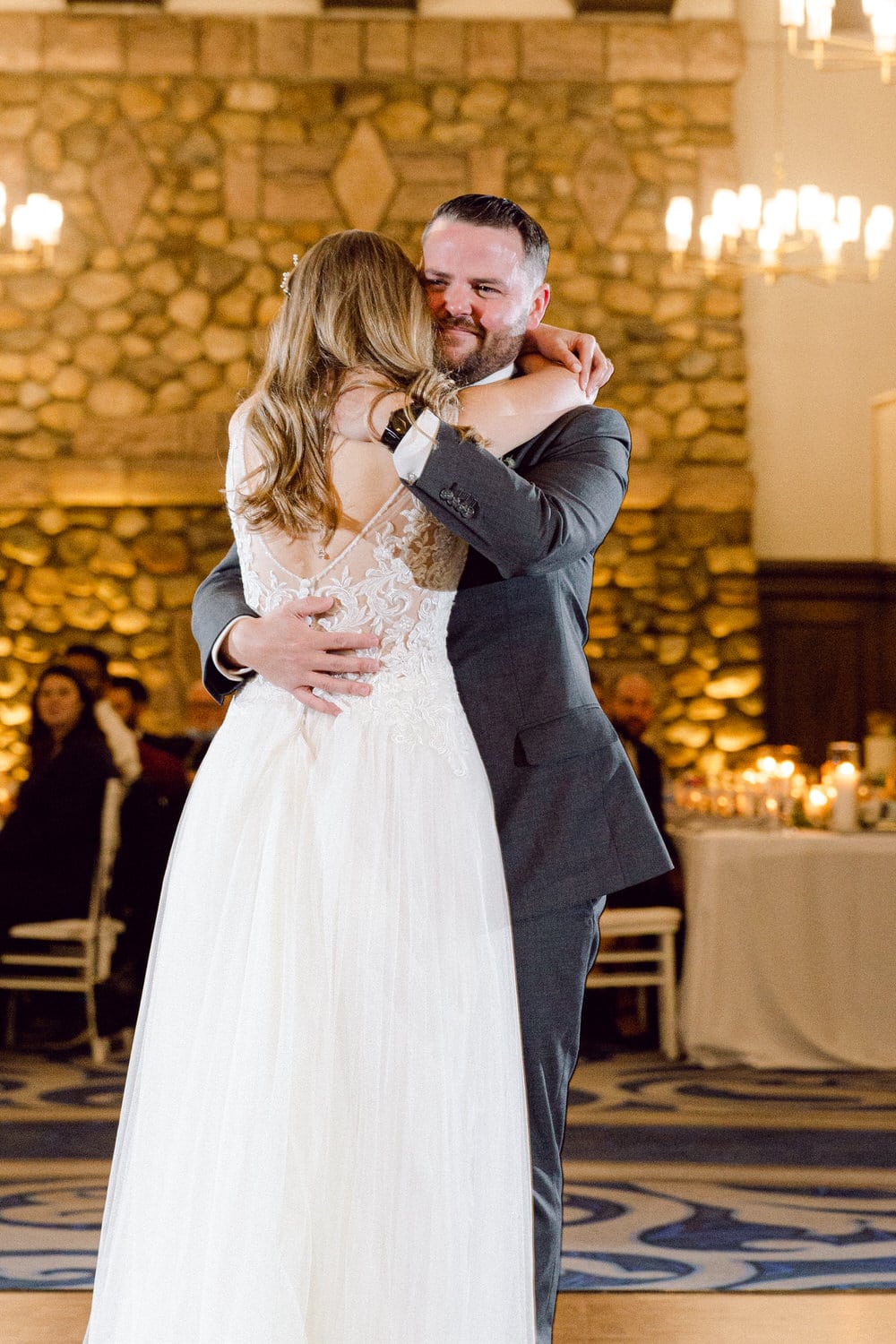 A bride and groom embrace while dancing together at their wedding reception, surrounded by softly lit decor and guests in the background.