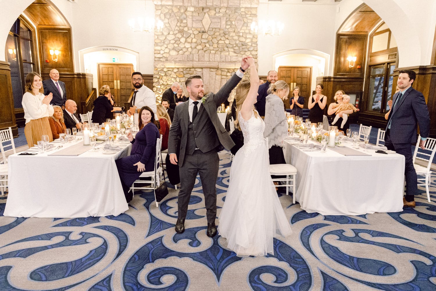 A couple joyfully dances together amid applauding guests at a wedding reception, with elegantly set tables and a beautiful stone fireplace in the background.