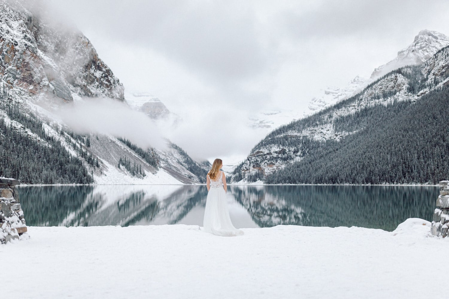 A woman in a white wedding dress stands on a snow-covered shore, gazing at a tranquil lake surrounded by snow-capped mountains and evergreen trees under a cloudy sky.