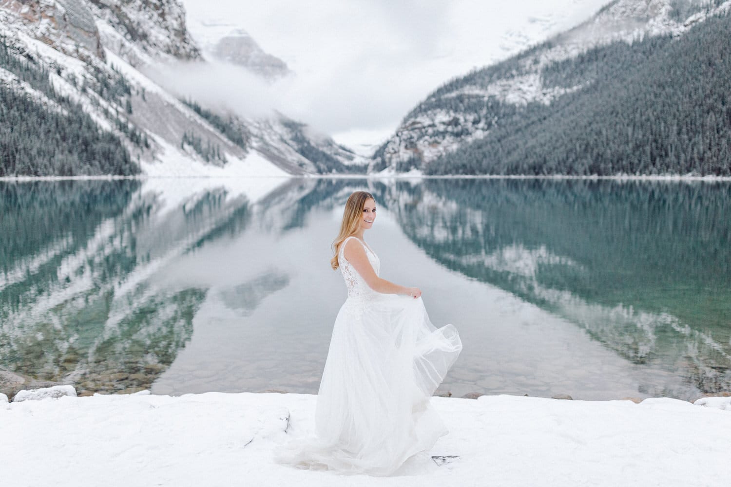 A woman in a flowing white dress stands on the snowy shore of a serene lake, surrounded by snow-covered mountains and mist, with her attire reflecting elegantly in the calm water.