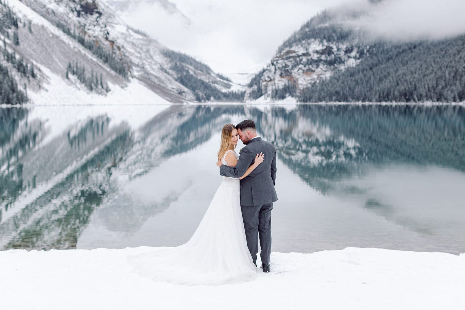 A couple embraces outdoors by a serene, snow-covered lake surrounded by mountains, reflecting their love in a tranquil winter landscape.