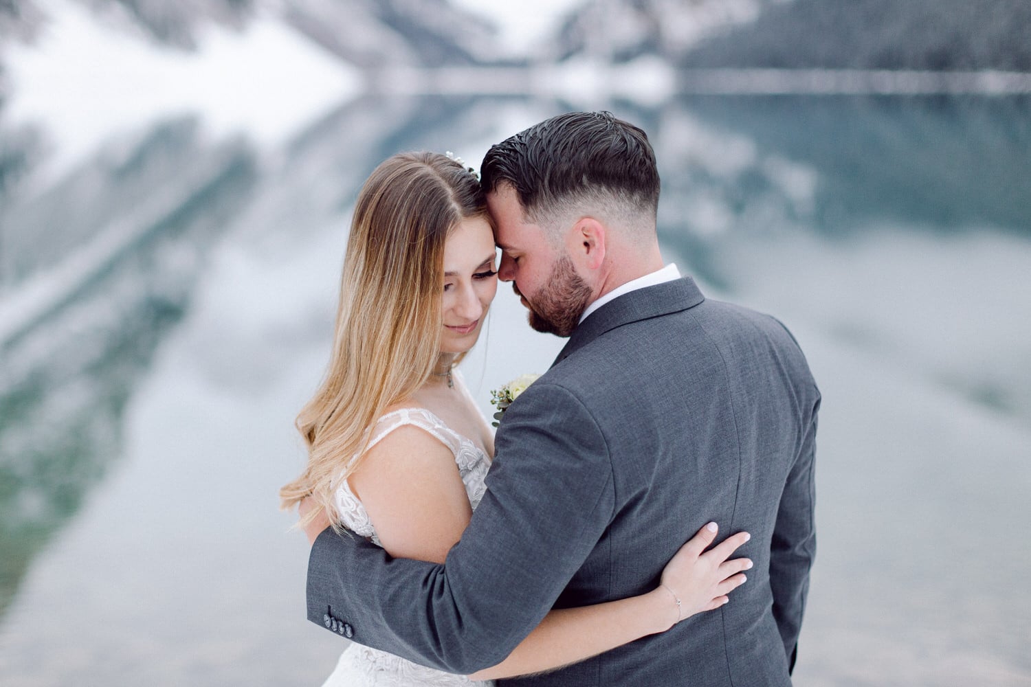 A bride and groom embrace tenderly by a serene mountain lake, reflecting a romantic atmosphere in a picturesque setting.