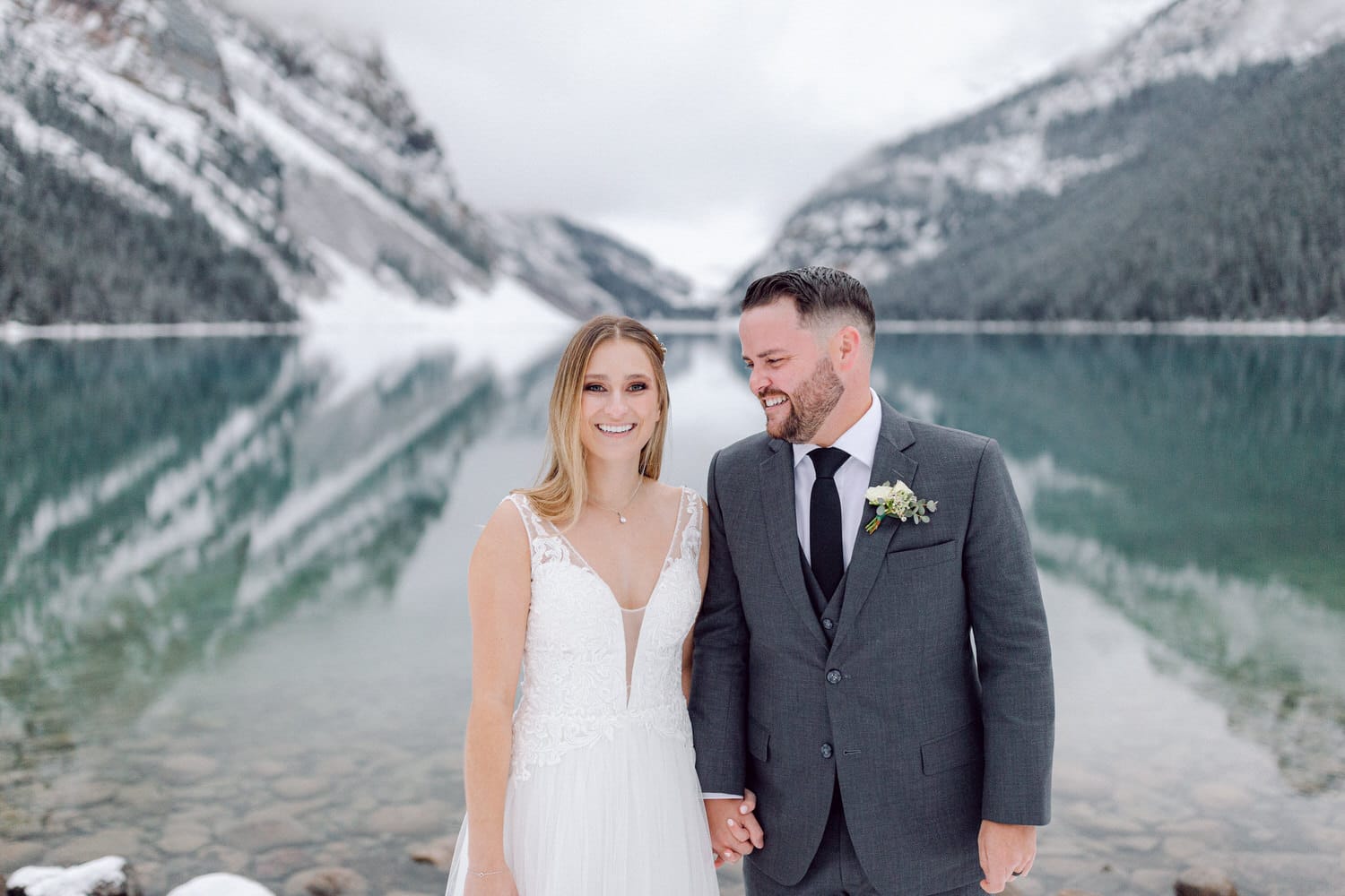 A joyful couple stands hand in hand by a serene, snow-capped lake, surrounded by majestic mountains and lush forest.