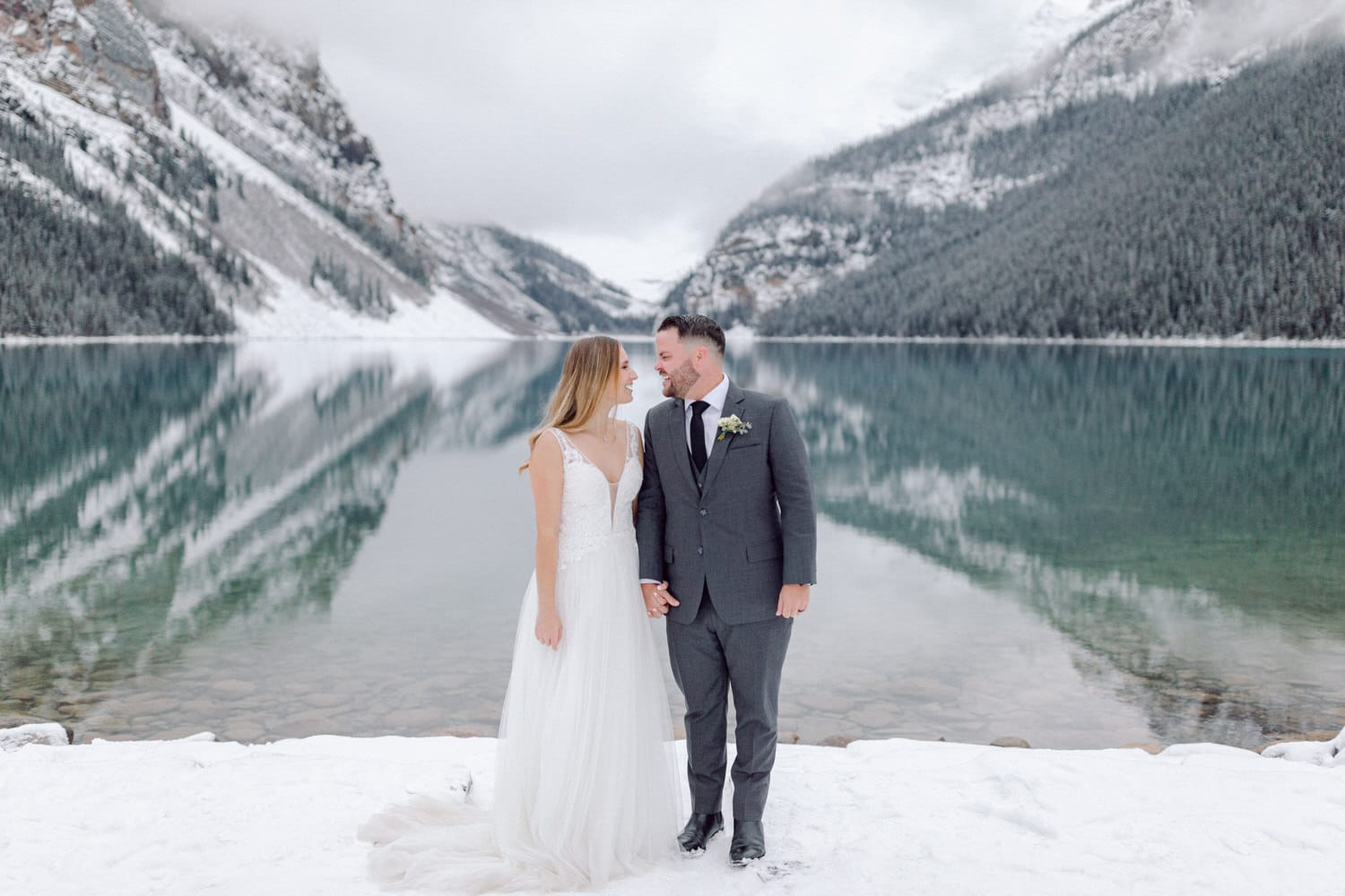 A bride and groom stand hand-in-hand by a snowy lake, surrounded by mountains and trees, with reflections in the calm water.