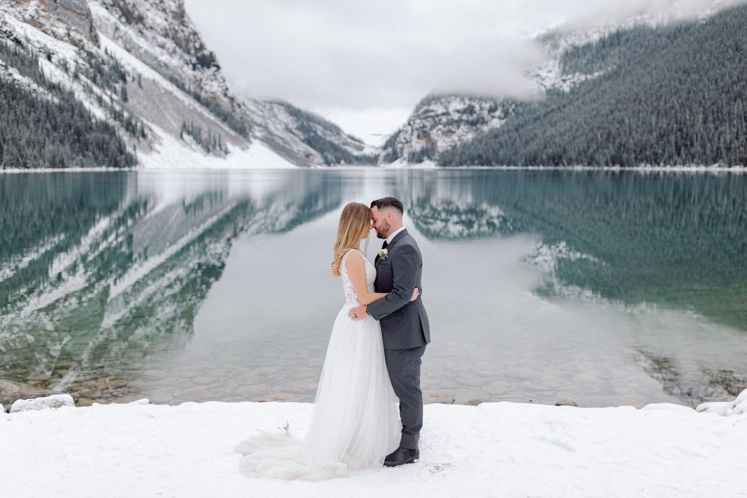 A couple embraces on the snowy shore of Lake Louise, with mountains and clouds reflected in the calm water behind them.