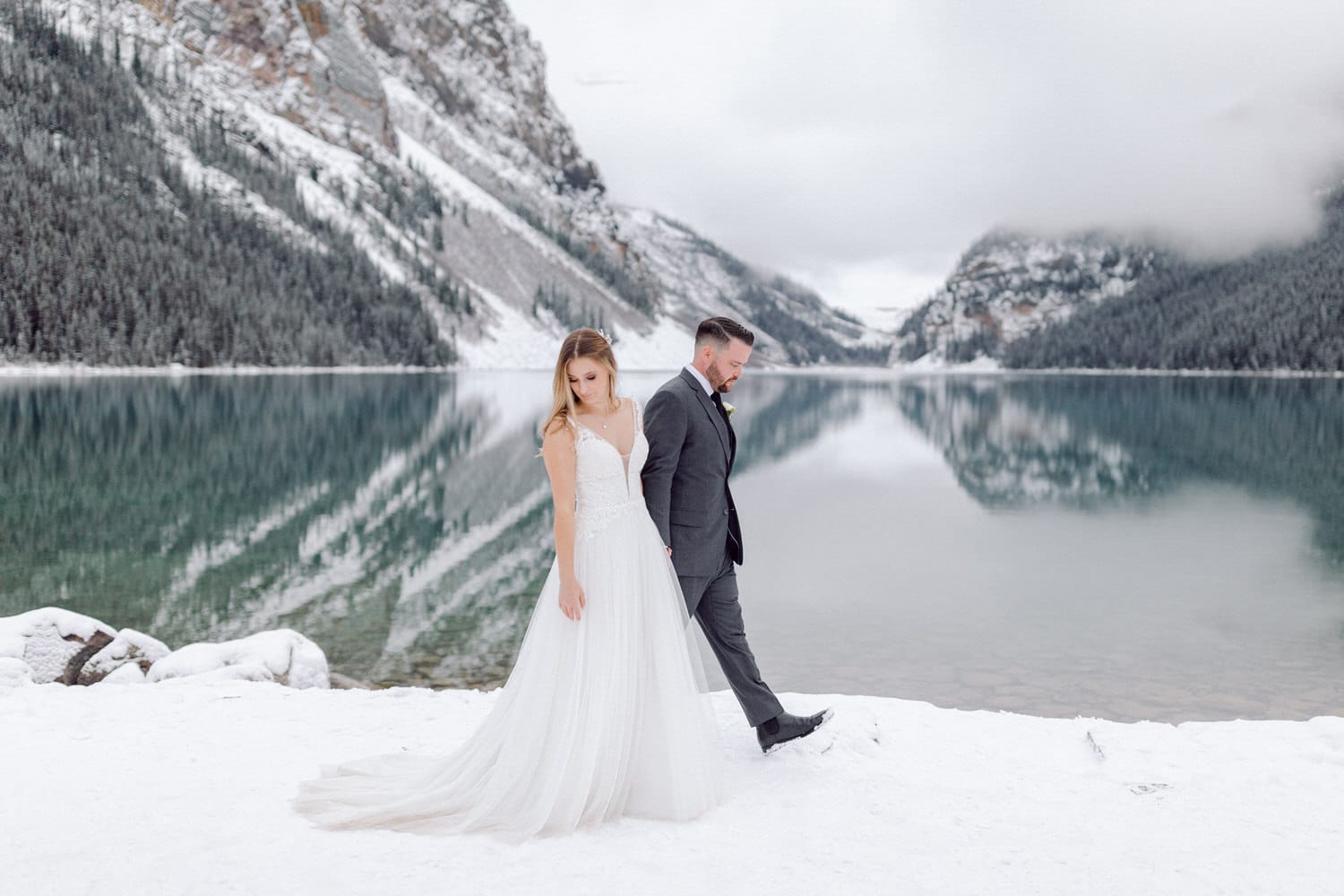 A couple walks hand in hand by a snow-covered lake surrounded by mountains, showcasing a romantic winter wedding scene.