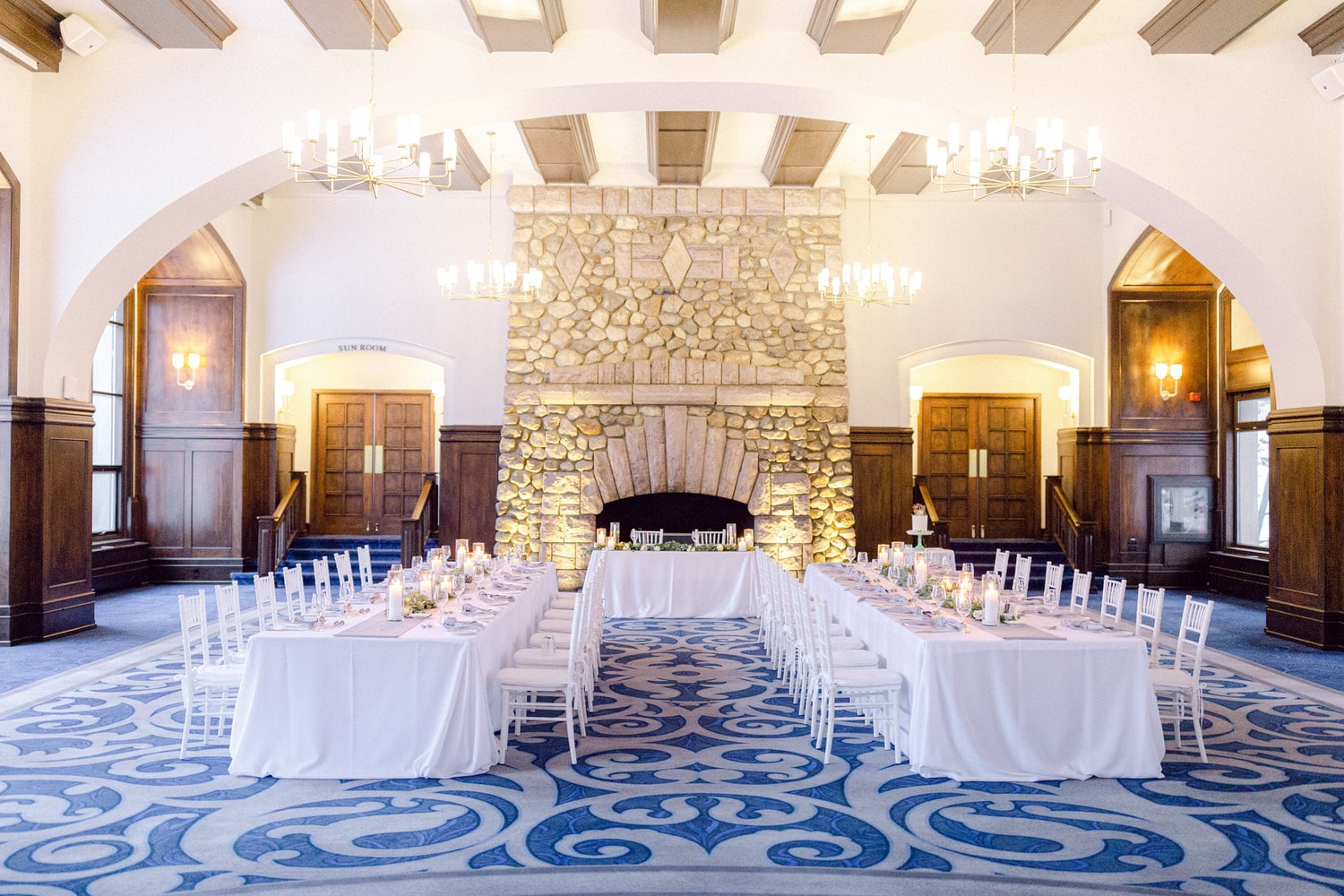 A beautifully arranged dining area with long tables adorned with white tablecloths, candles, and decor, featuring a stunning stone fireplace in the background.