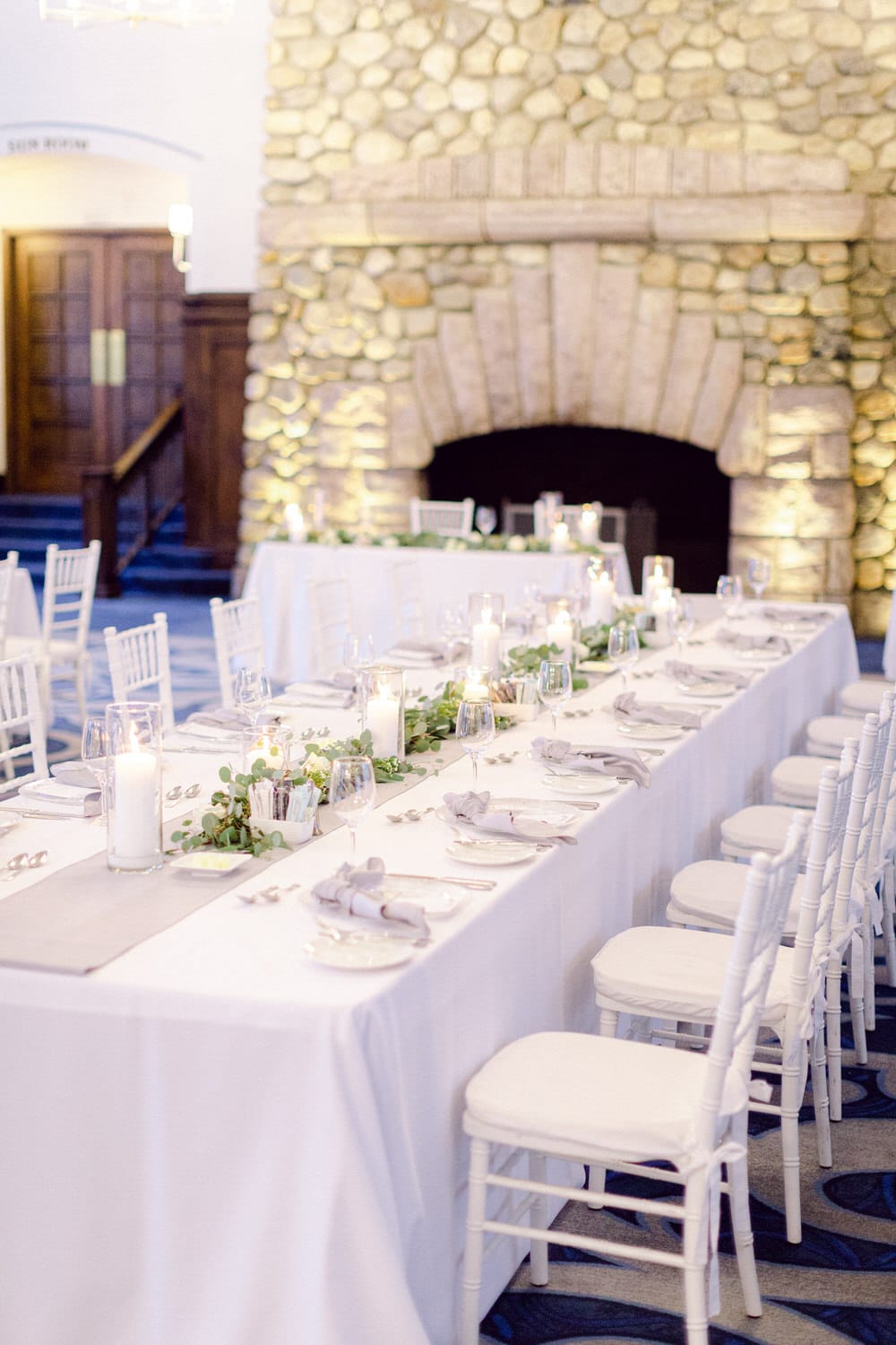 A beautifully arranged long dining table adorned with white linens, green foliage, candles, and elegant tableware, set against a backdrop of a stone fireplace and wooden details.