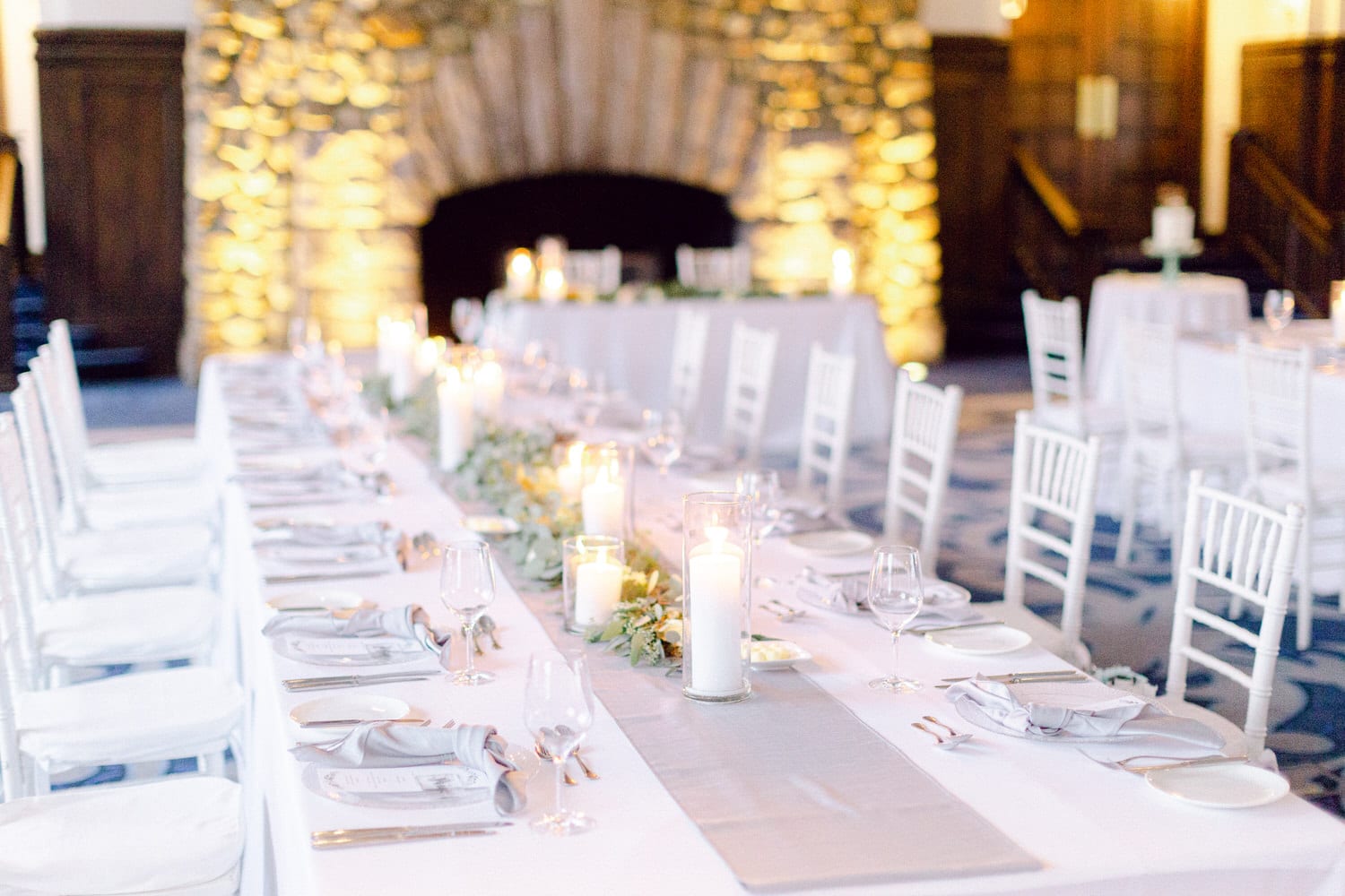 A beautifully arranged long dining table with white chairs, adorned with candles and greenery, set against a backdrop of a stone fireplace.