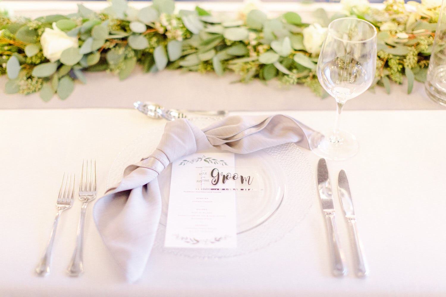 A beautifully arranged wedding table featuring a place setting with a menu card labeled 'groom', delicate silverware, and a soft grey napkin, complemented by greenery in the background.