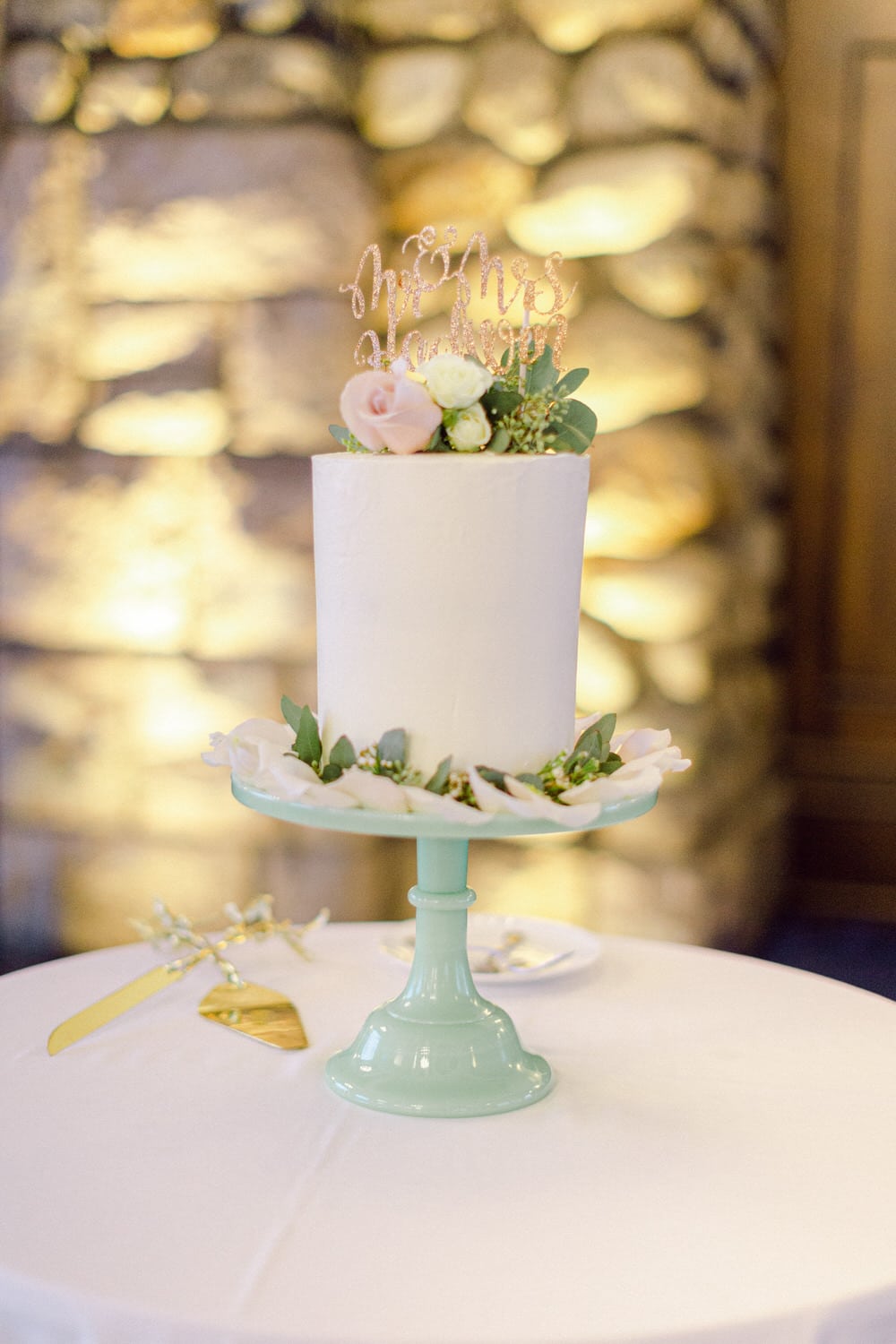 A beautifully decorated white wedding cake with floral accents on a mint-green cake stand, topped with a sparkling "Mr. & Mrs." sign, set against a softly lit background.