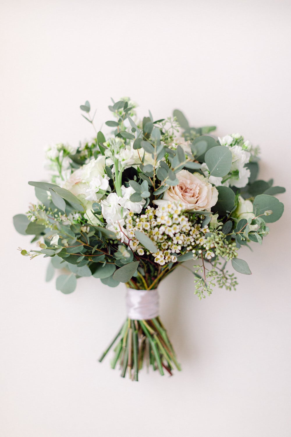 A delicate bouquet featuring soft peach roses, white flowers, and lush greenery, elegantly wrapped in a satin ribbon against a neutral background.