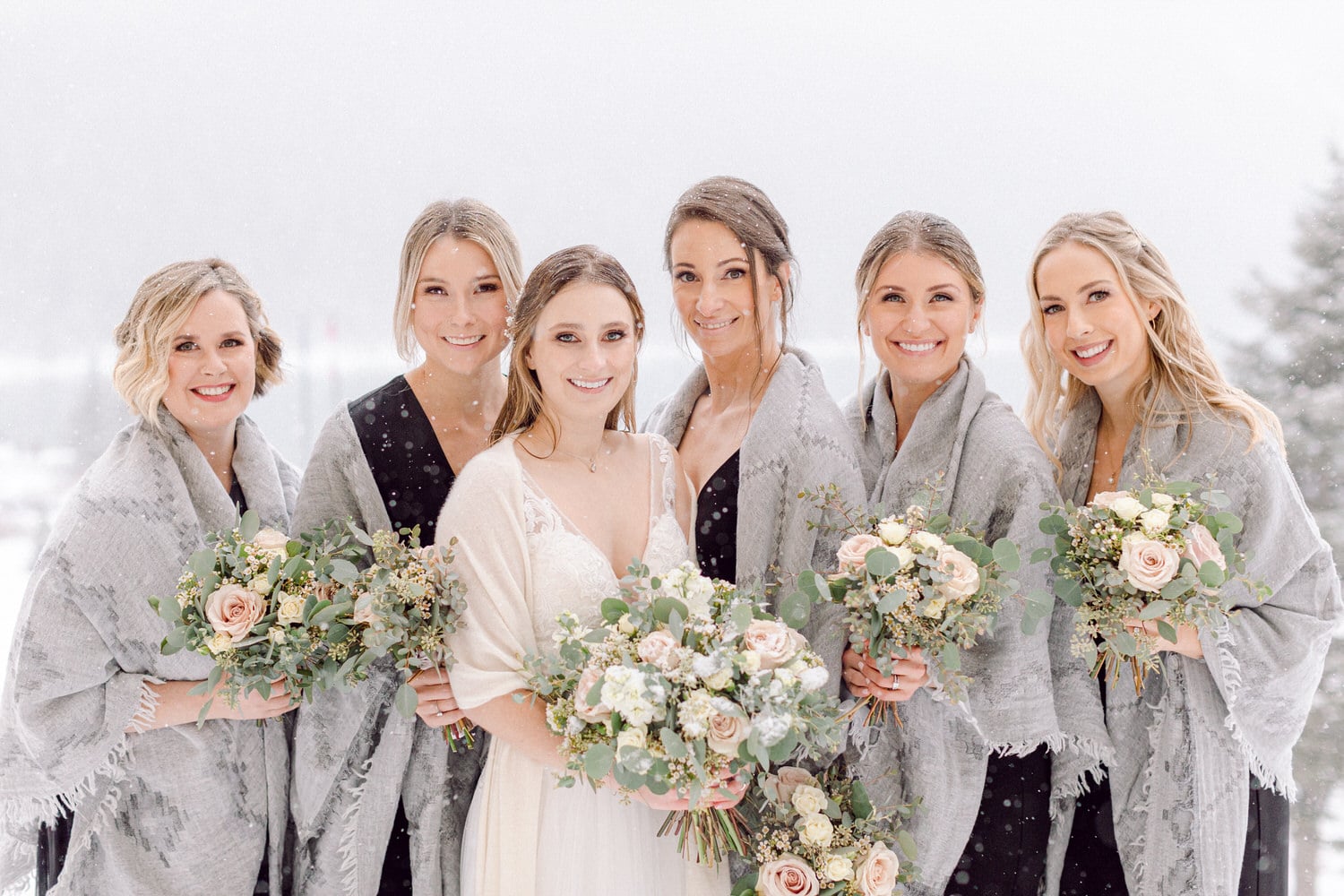 A bride and her five bridesmaids joyfully pose together in a snowy outdoor setting, all wearing elegant shawls and holding beautiful floral bouquets.