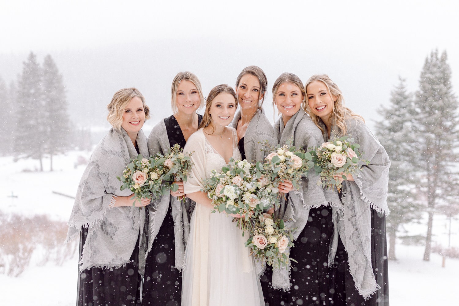 A bride surrounded by her six bridesmaids, all smiling amidst falling snow, holding bouquets of flowers, wearing matching shawls.