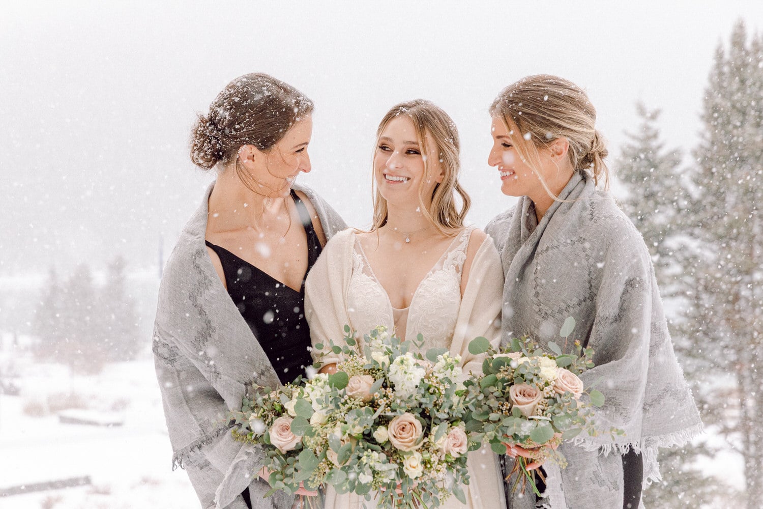 Bridesmaids in elegant shawls smiling and holding floral bouquets amidst a snowy backdrop.