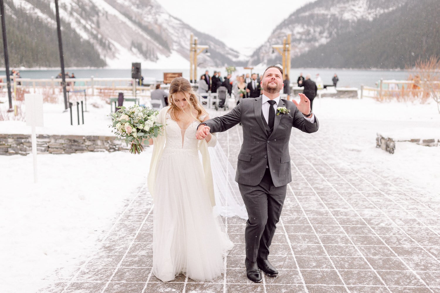 A joyful couple walks hand-in-hand through a snowy outdoor wedding venue, with mountains and guests in the background.