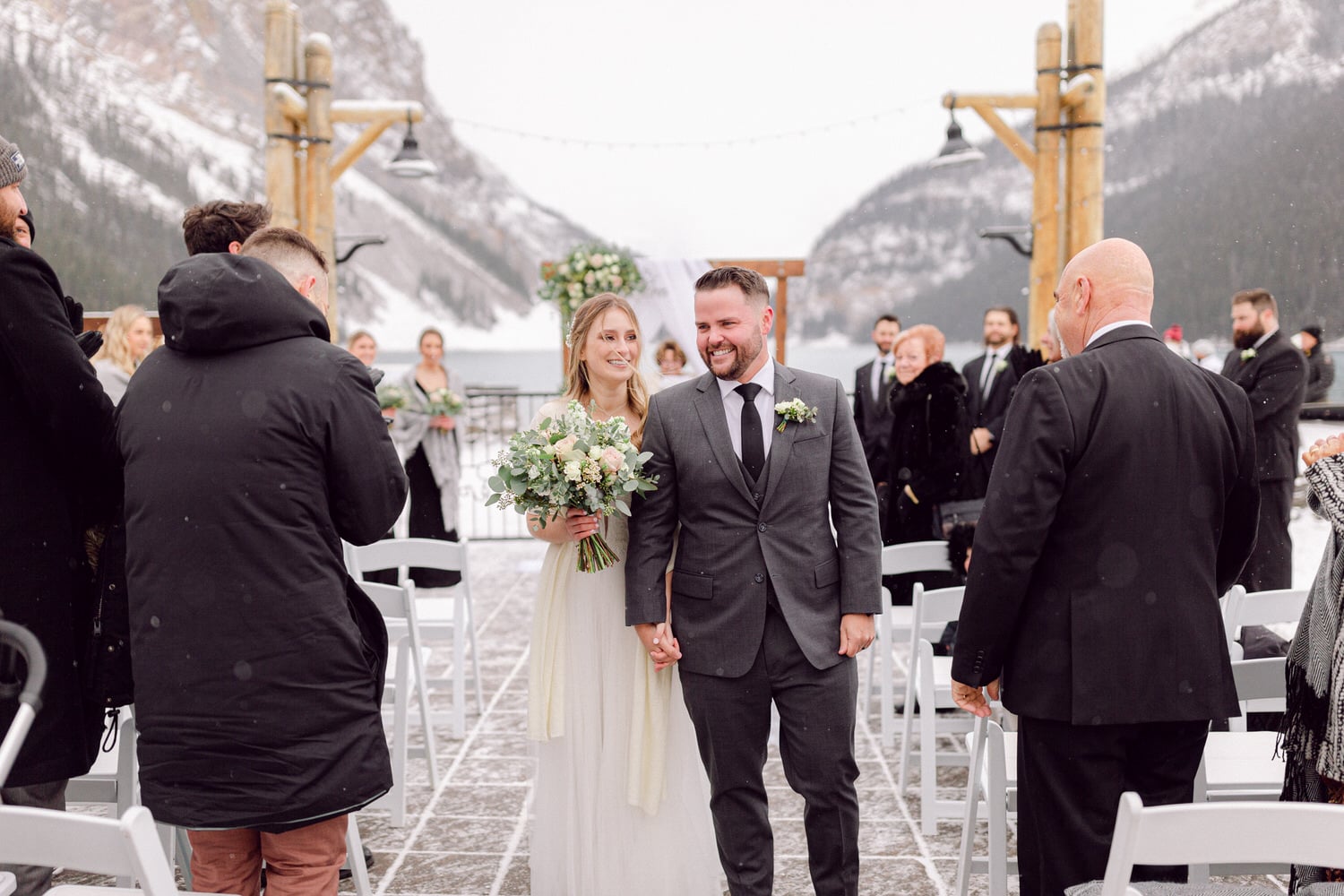 A newlywed couple joyfully walks hand in hand down an outdoor aisle surrounded by snow-covered mountains and guests celebrating their union.