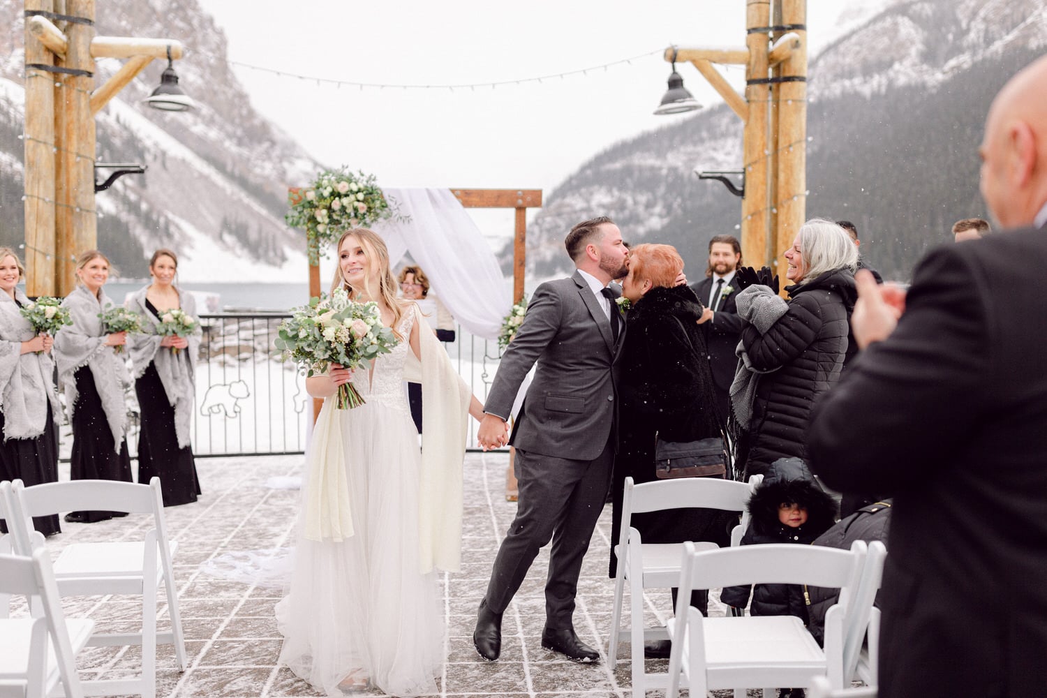 A couple shares a kiss after exchanging vows in a winter wedding ceremony, surrounded by family and friends in a picturesque snowy landscape.