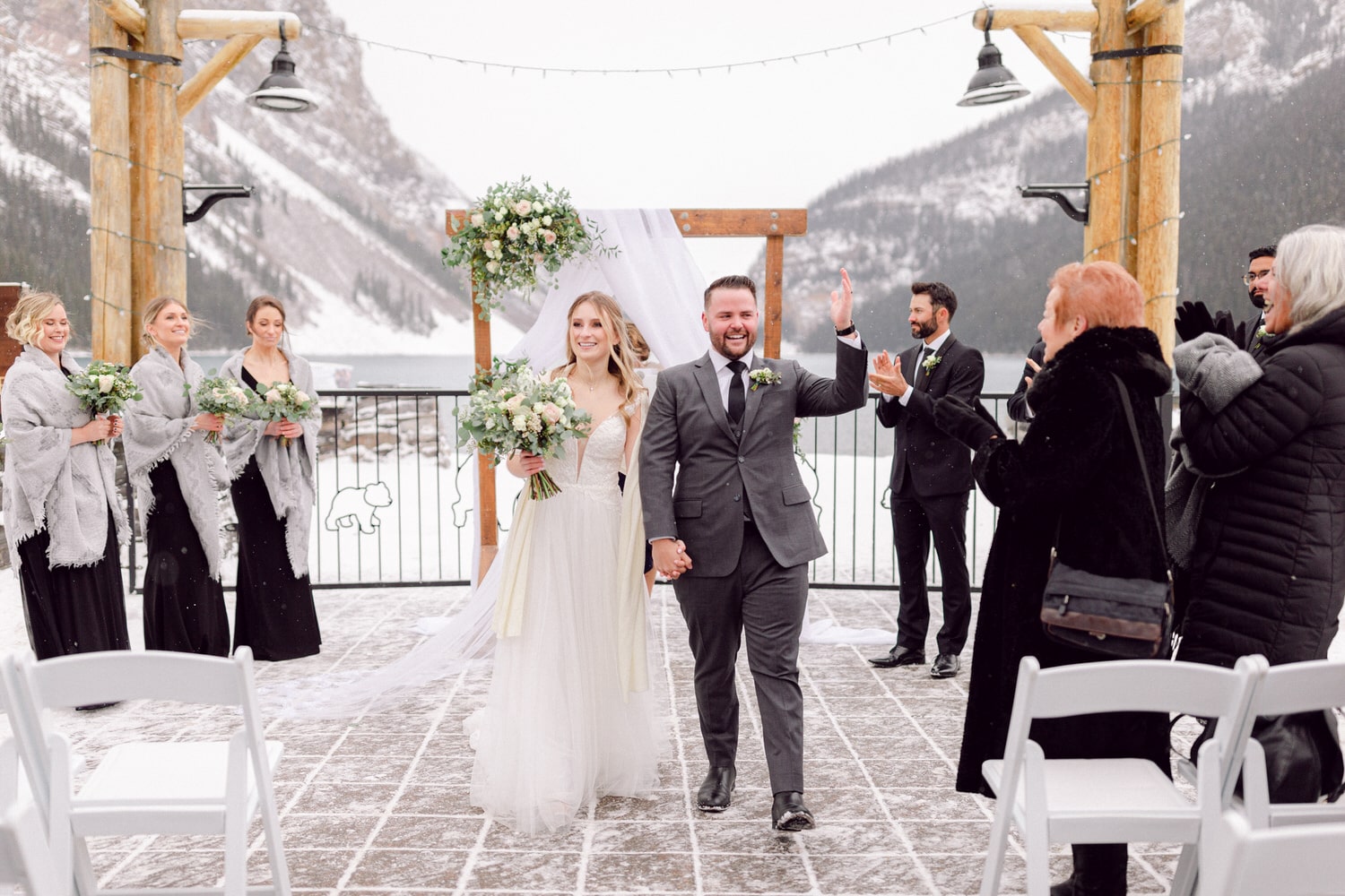 A joyful couple walking hand in hand after their wedding ceremony, surrounded by snow and mountains, with guests applauding and bridesmaids holding bouquets.