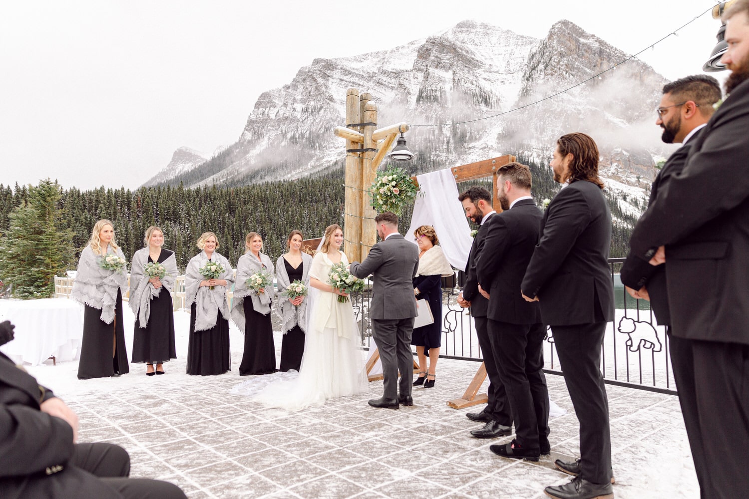 A bride and groom exchange vows during a winter wedding ceremony surrounded by guests, with snow-capped mountains in the background.