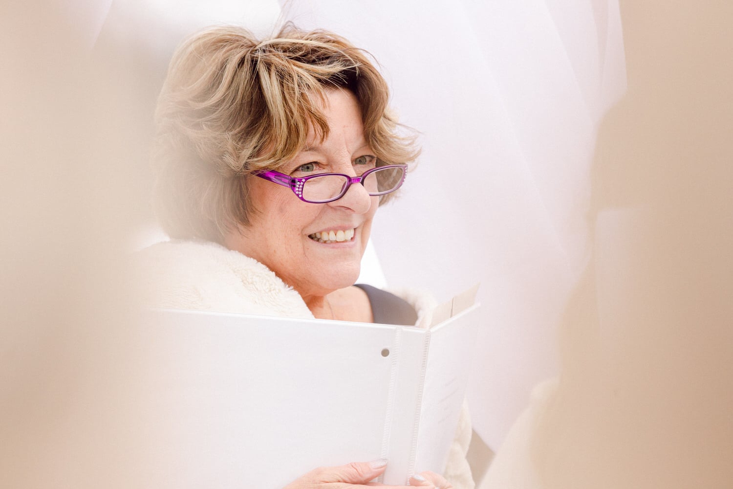 A smiling woman with short, wavy hair and purple glasses holds a binder, dressed in a cozy white shawl, sitting in a softly lit setting.