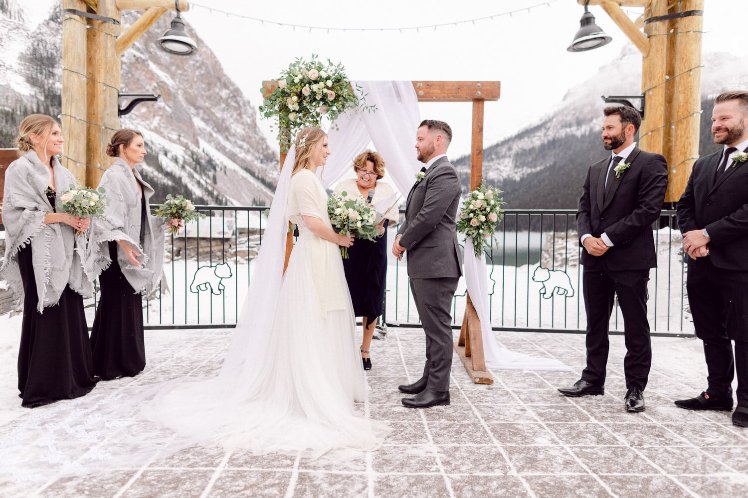 A bride and groom exchange vows under a floral arch amidst snowy mountains, surrounded by wedding party members in elegant attire.