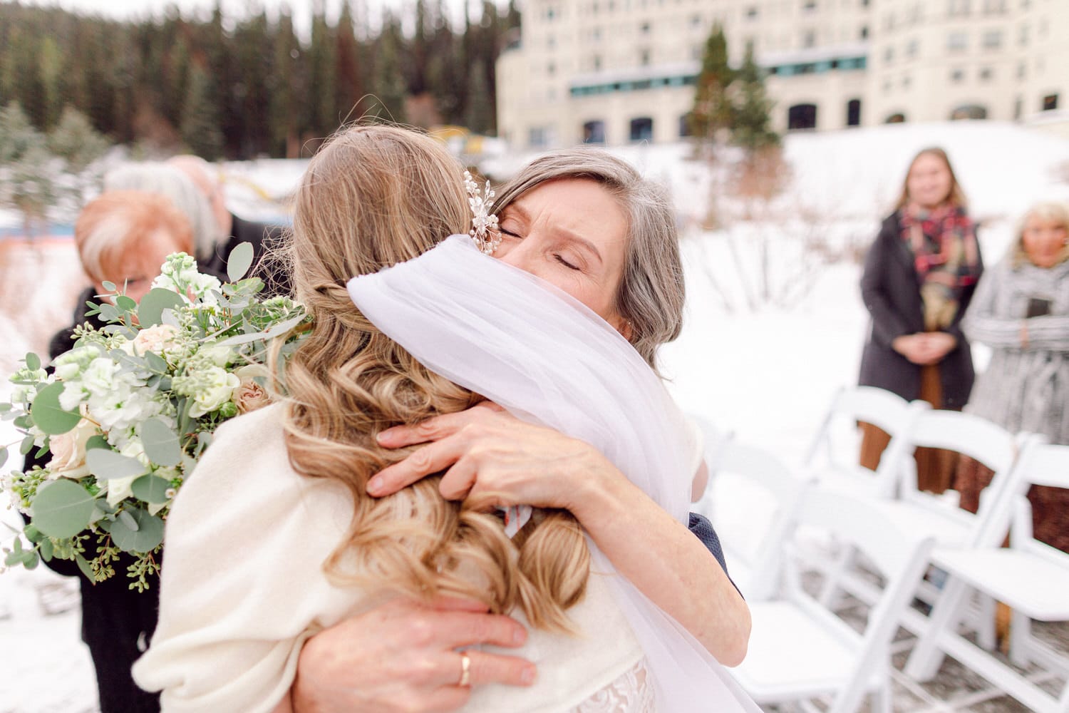 A bride is joyfully hugging her mother in a snowy outdoor setting, surrounded by guests and a beautiful winter backdrop.