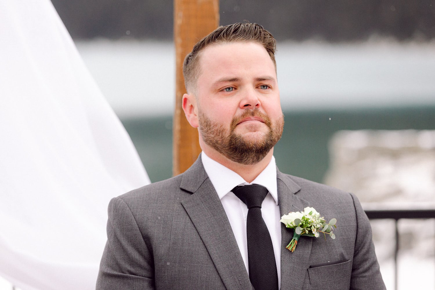 A groom in a gray suit with a black tie and floral boutonniere, standing solemnly during an outdoor winter wedding against a snowy backdrop.