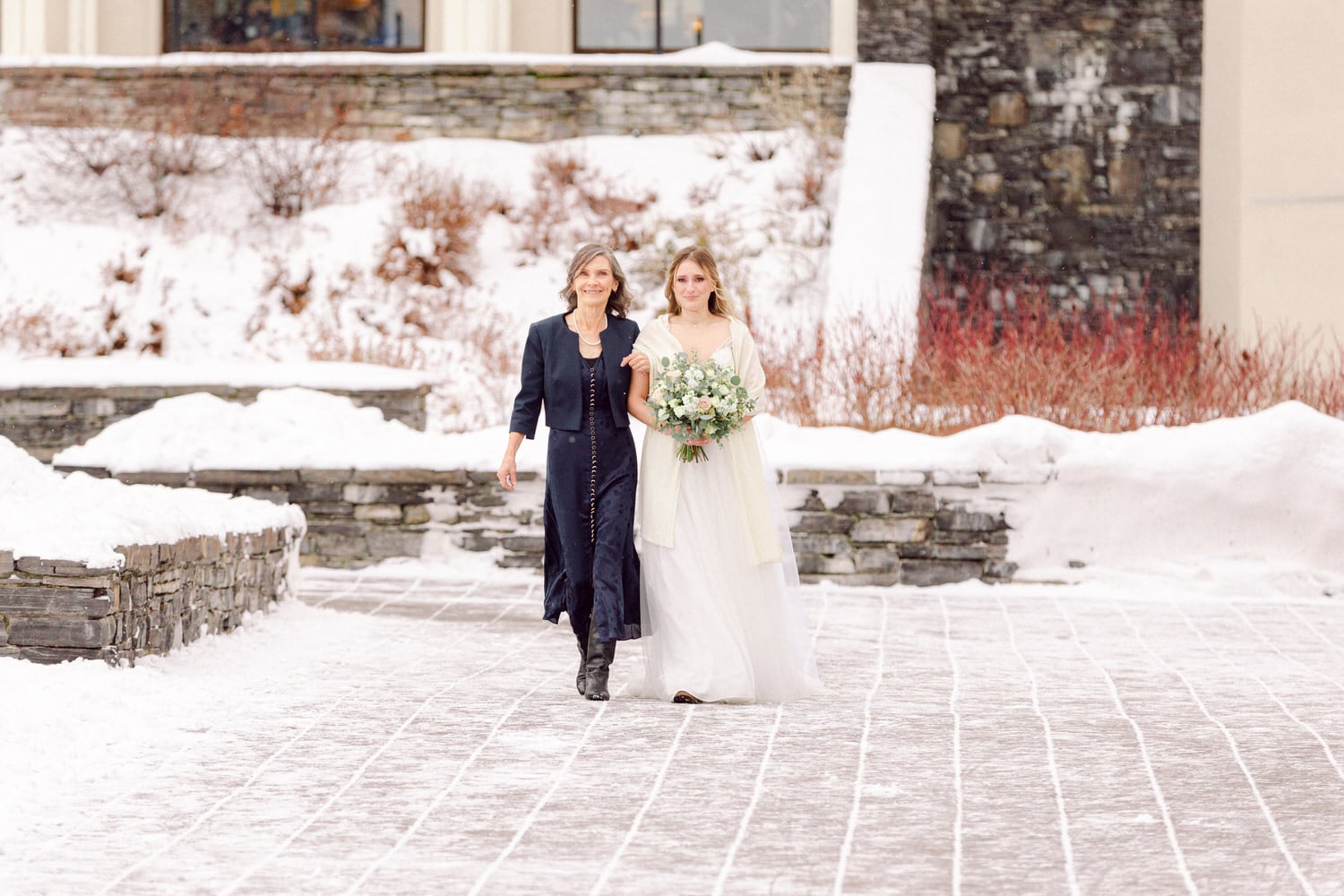 A bride and her companion walking together on a snowy pathway, surrounded by winter scenery, showcasing the beauty of a serene wedding day.