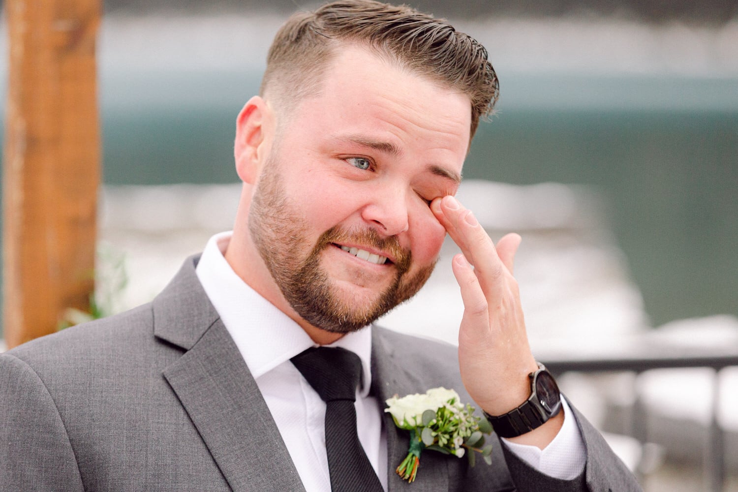 A groom brushes away tears during a heartfelt wedding ceremony, wearing a grey suit and boutonnière, capturing the essence of love and emotion.