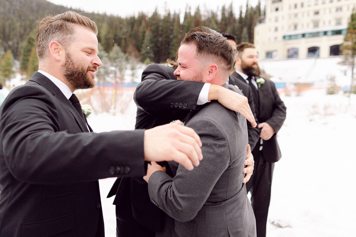 A group of well-dressed men sharing a heartfelt embrace in a snowy landscape, with trees and a building in the background.