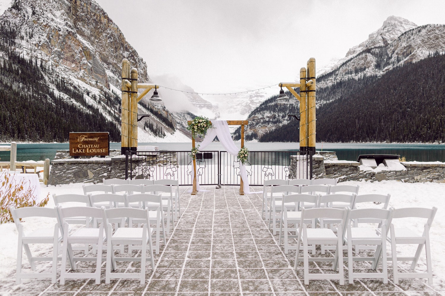 A serene wedding venue with white chairs arranged facing an ornate altar, surrounded by snow-covered mountains and the tranquil waters of Lake Louise.