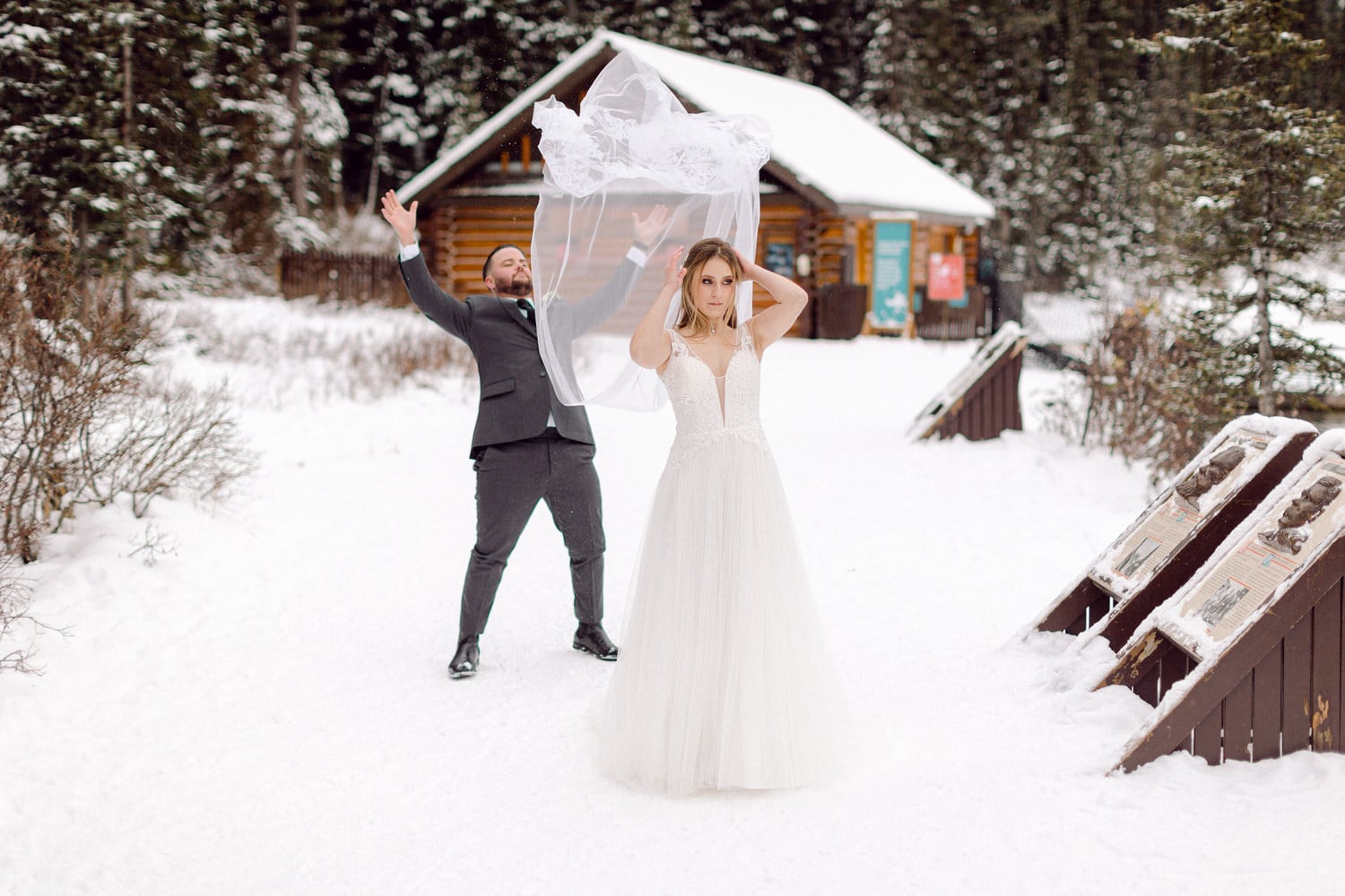 A bride in a flowing white gown holds her veil while a man in a suit playfully throws it into the snow-covered landscape.