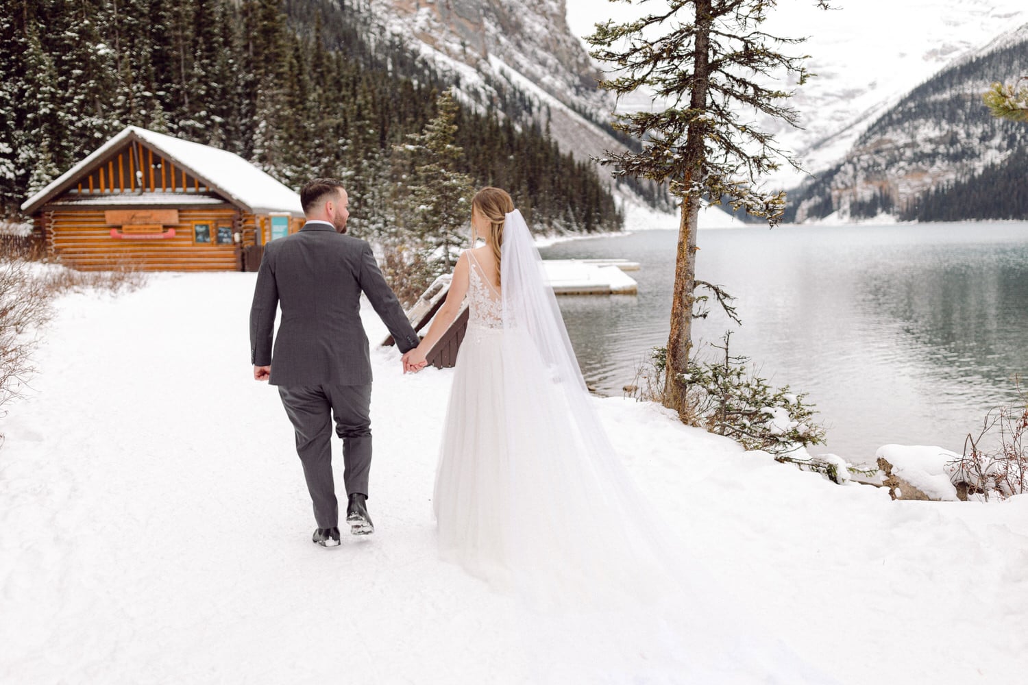 A couple walks hand in hand along a snowy path by a lake, surrounded by evergreen trees and a log cabin in the background.