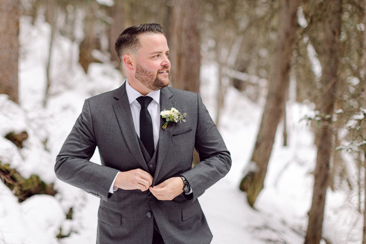 A groom in a gray suit with a boutonniere adjusts his jacket while standing in a snowy forest backdrop.